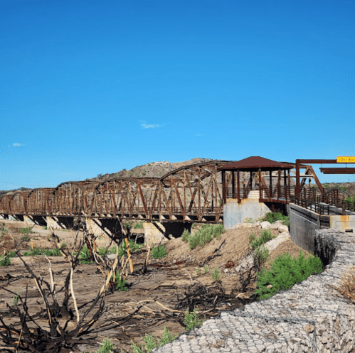 A wooden bridge spans a dry riverbed, surrounded by sparse vegetation and a clear blue sky.